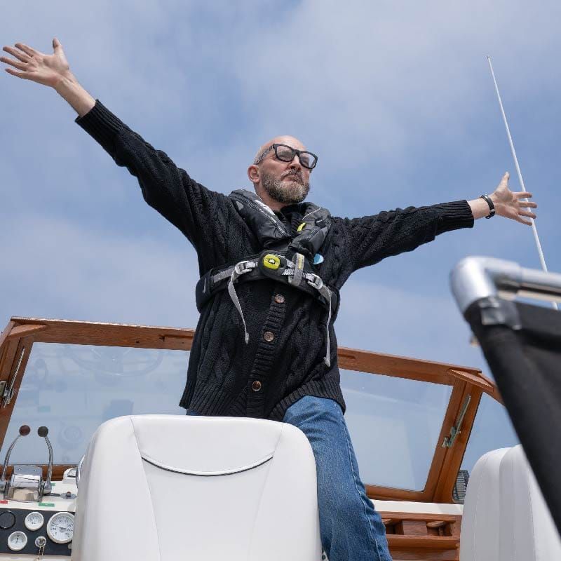 Man with open arms on a boat, wearing life vest. Blue sky, water.