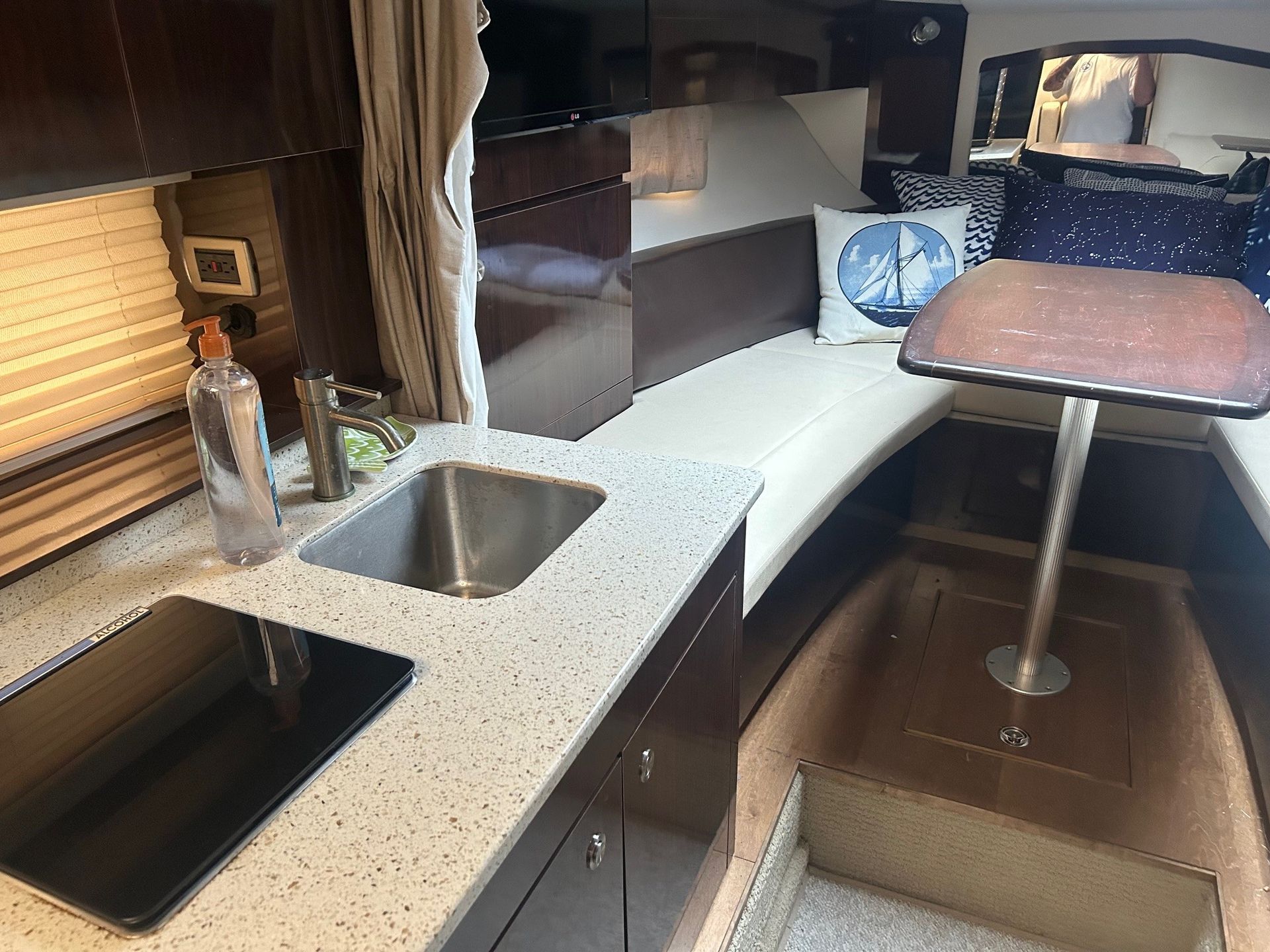 Interior of a boat cabin with a galley, seating, and bed. White countertops, dark wood, and neutral tones.
