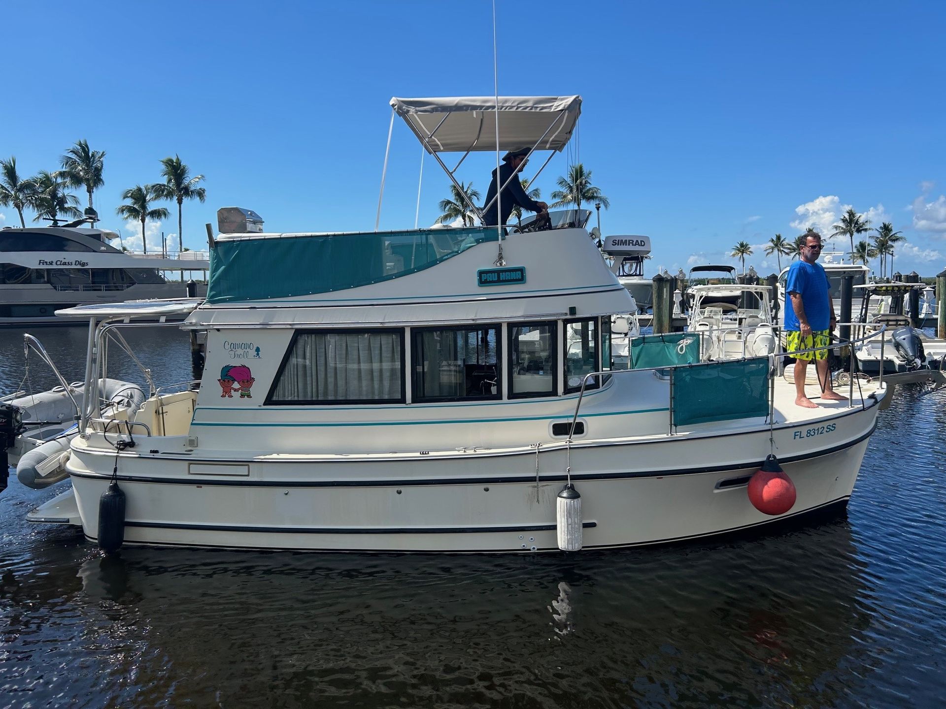 White and teal houseboat in water with a man, parked at a dock with palm trees and blue sky.