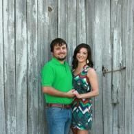 Couple smiling and holding hands in front of a weathered wooden wall.