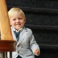 Smiling child in a light gray suit sitting on carpeted stairs near a wooden railing