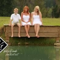 Three people in white outfits sit on a wooden dock by a calm green lake.