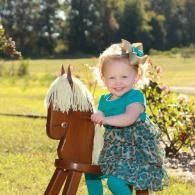 Smiling child in teal dress standing beside a brown wooden hobby horse outdoors