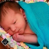 Sleeping newborn wrapped in a turquoise blanket on a colorful floral blanket, with its hand near the mouth