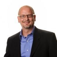 Smiling man in glasses wearing a black blazer and blue shirt on a white background
