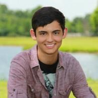 Smiling person in a pink shirt sitting outdoors by a lake with green trees in the background