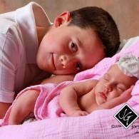 Young boy resting beside a sleeping baby on a pink blanket,  lying close together, him looking at the camera