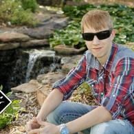 Person in sunglasses and red plaid shirt sitting outdoors by a small waterfall and rocks