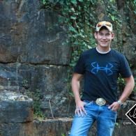 Person standing against a rocky wall in a black shirt, jeans, and cap, outdoors