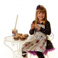 Young child in a polka-dot dress sitting at a small table with tea and pastries, smiling at the camera