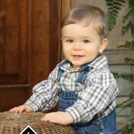 Toddler in plaid shirt and denim overalls, smiling and leaning on a woven chair indoors