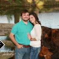 Couple posing by a lake, with a blanket hanging on a wooden fence in the background