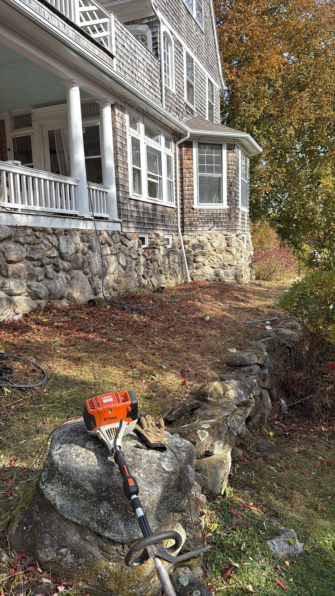 String trimmer on rock in front of a stone house with porch and autumn trees.
