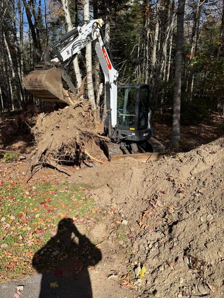 Bobcat excavator digs earth, trees in background, autumn setting. Shadow of person in foreground.