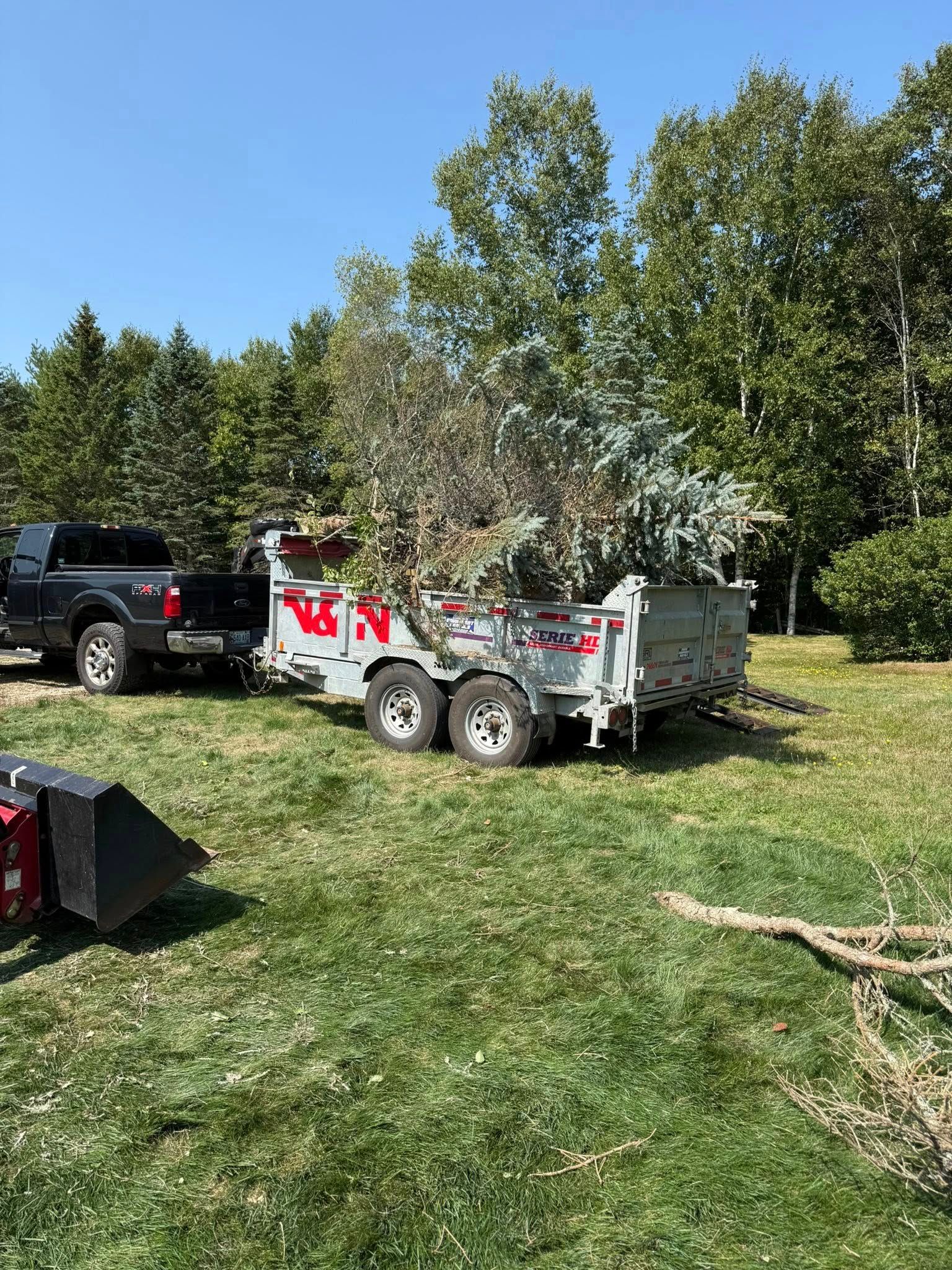 Black truck towing a gray trailer loaded with tree branches on a grassy lawn under a blue sky.