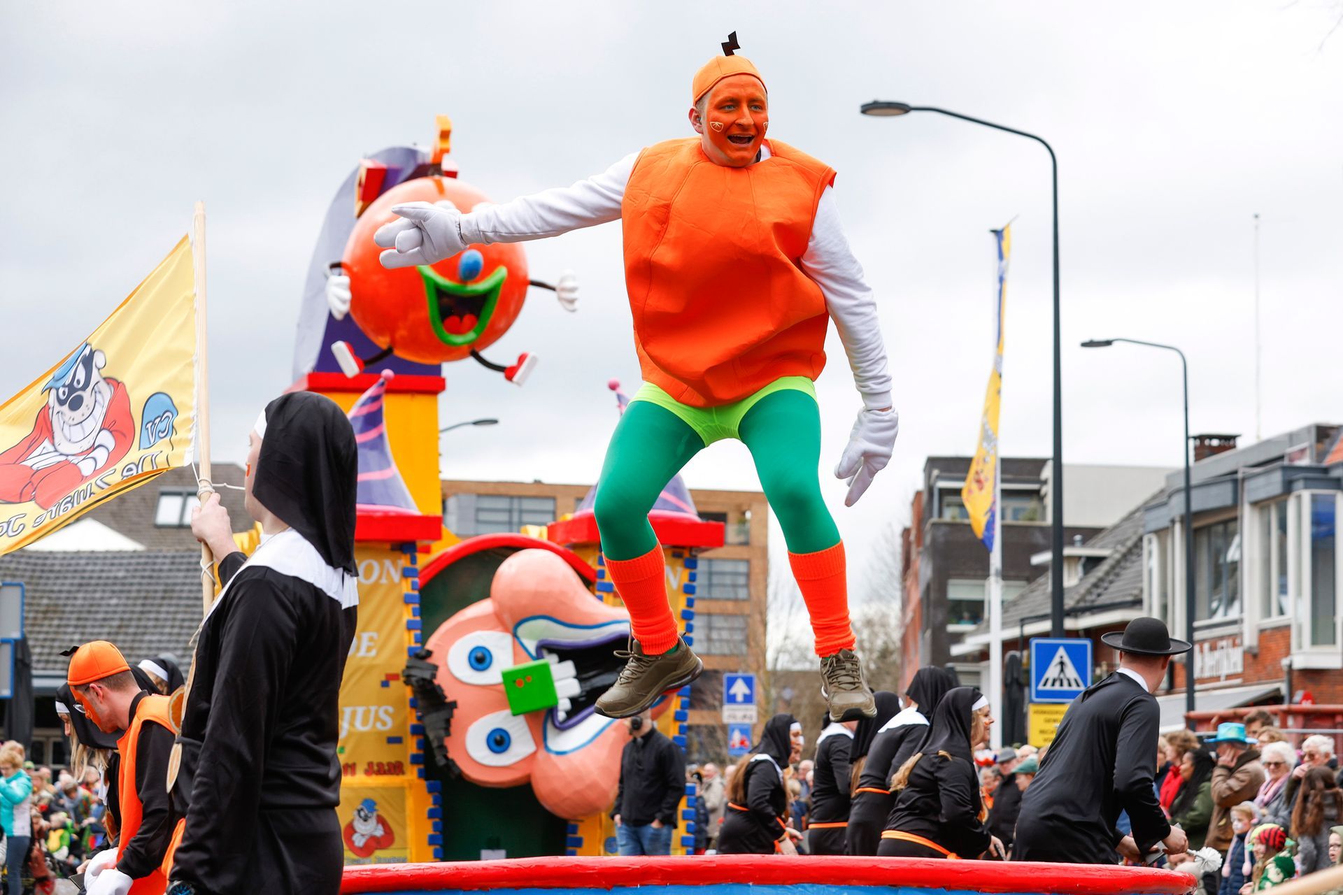 Een man in een oranje kostuum springt in de lucht op een praalwagen tijdens een parade.
