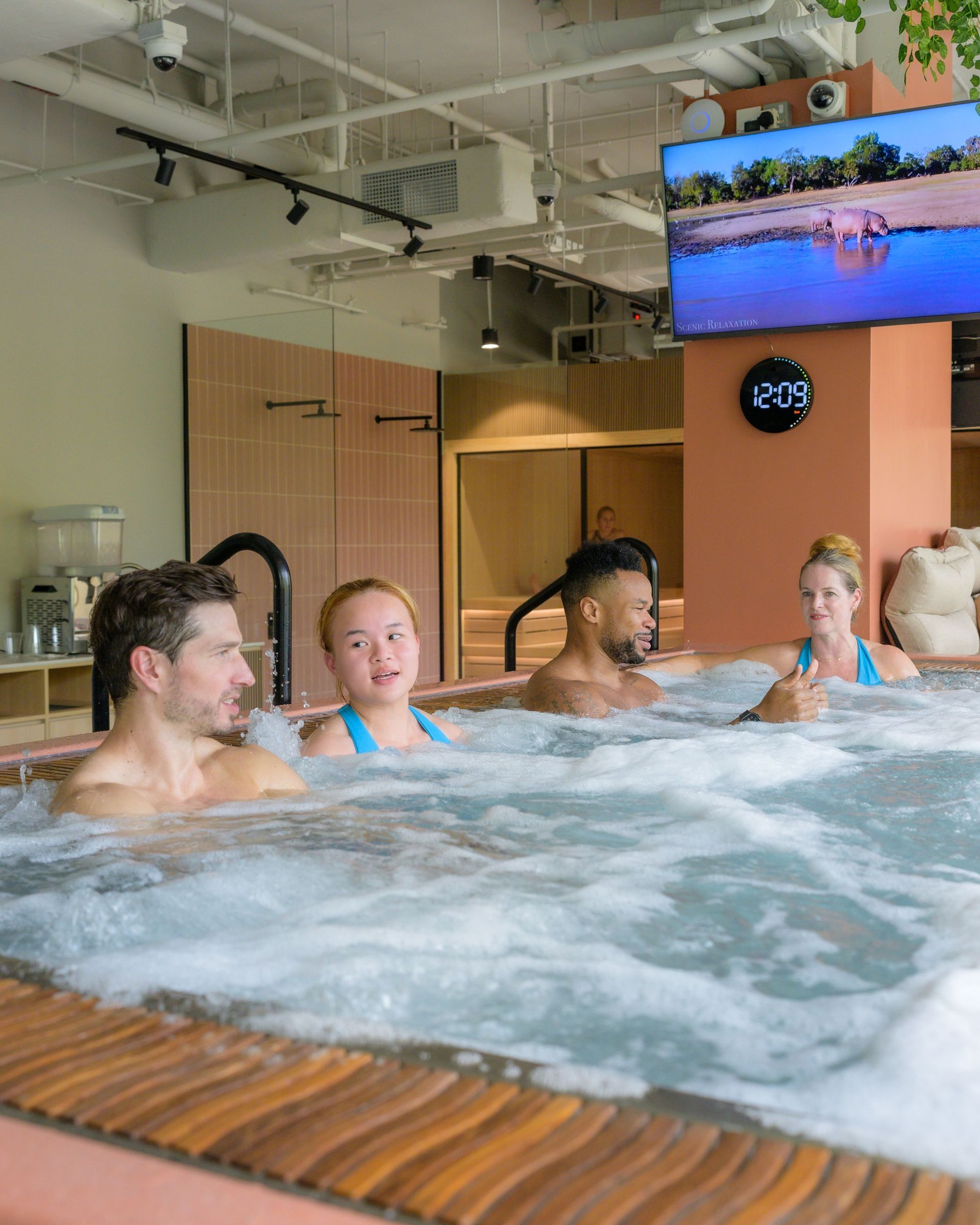Four people smiling in a spa pool, steam rising. Large window in the background.