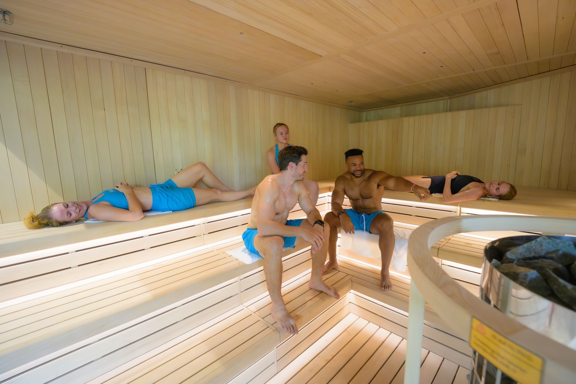 Woman sitting on a sauna bench, eyes closed, black swimsuit. Wooden interior with soft lighting.