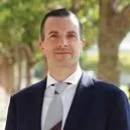Man in dark suit and tie, smiling outdoors with a blurred tree background.