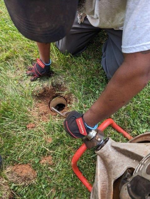 A man is kneeling down in the grass using a tool to fix a hole in the ground.