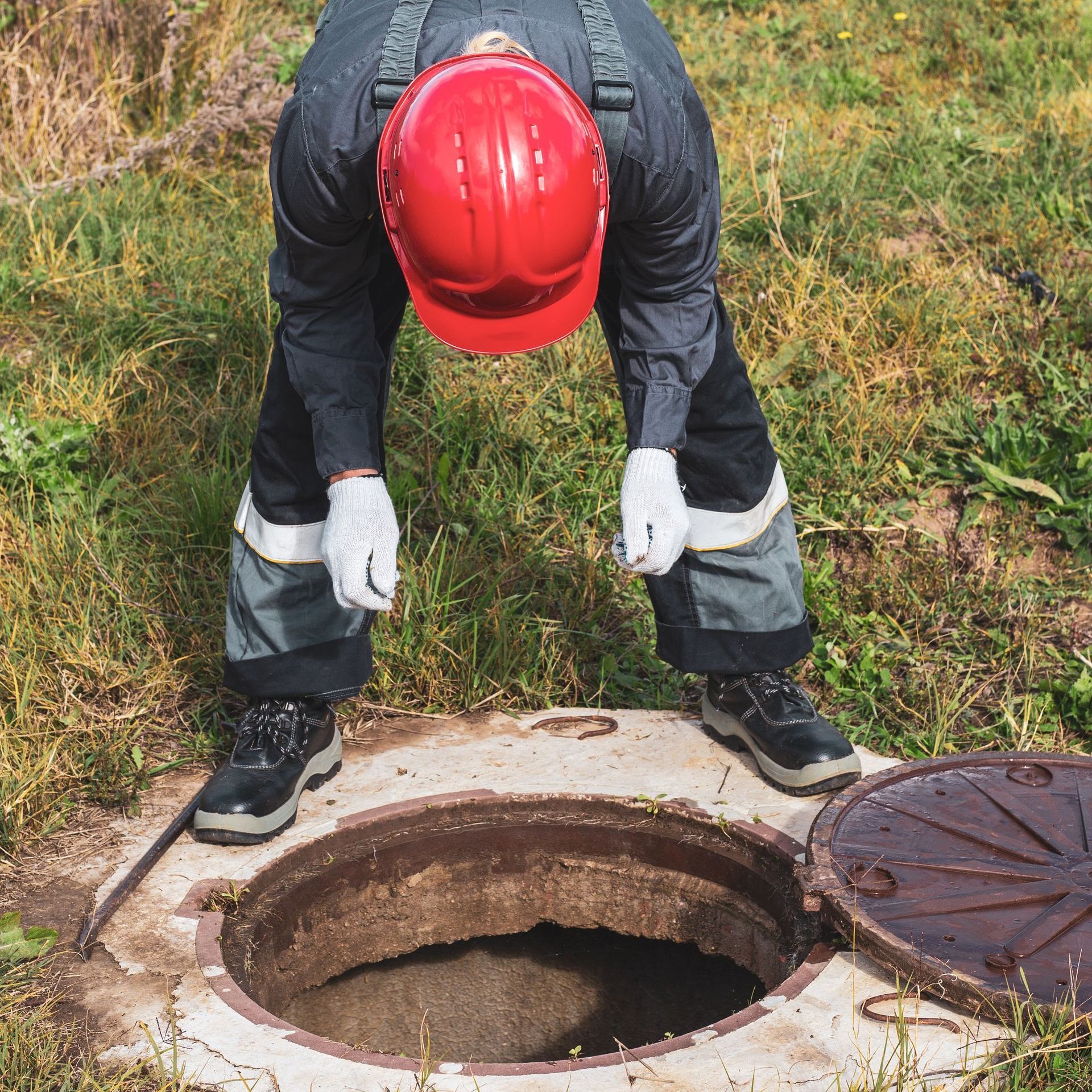 A man wearing a red hard hat is looking into a manhole cover.