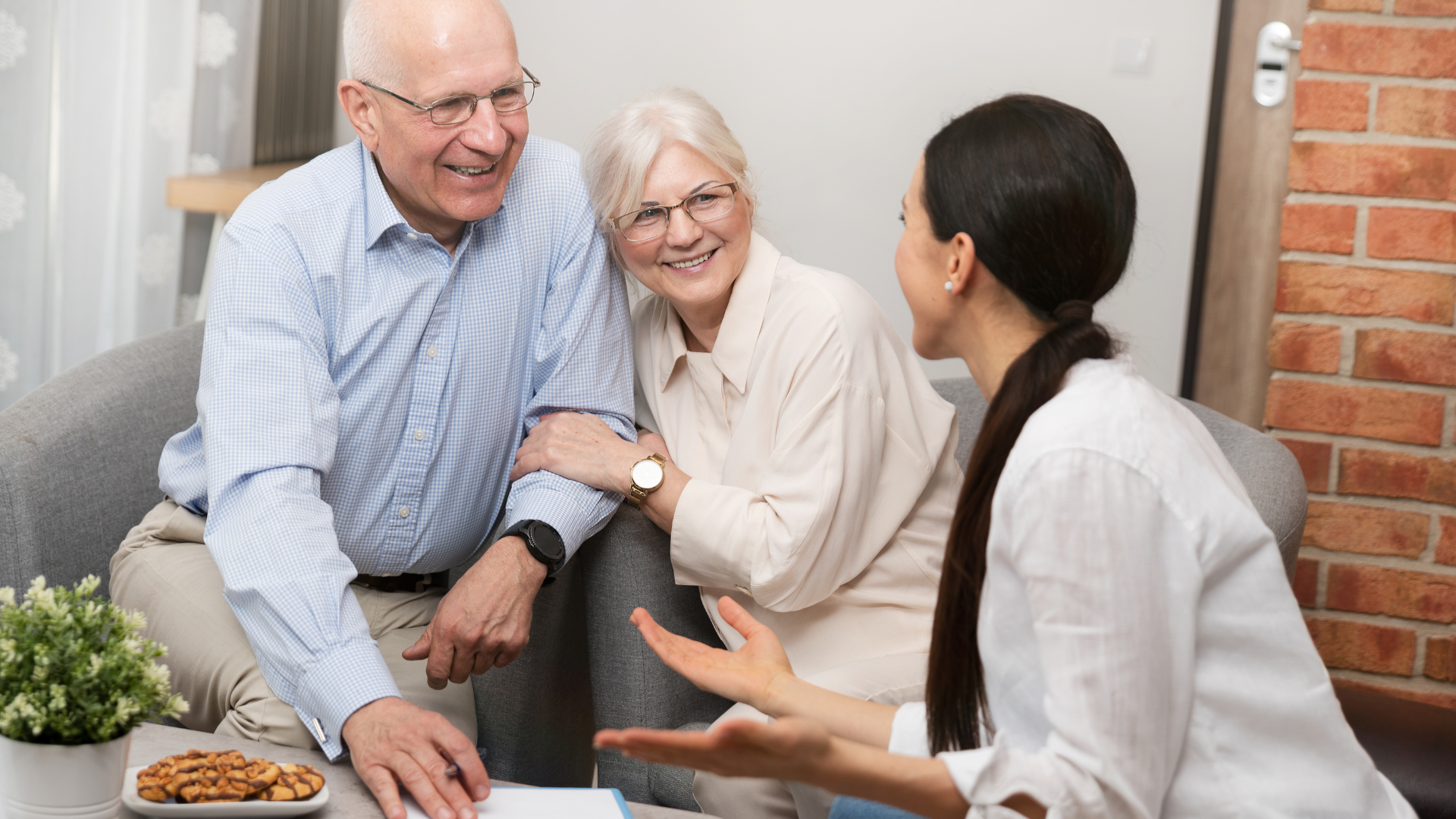 An elderly couple is sitting on a couch talking to a woman.