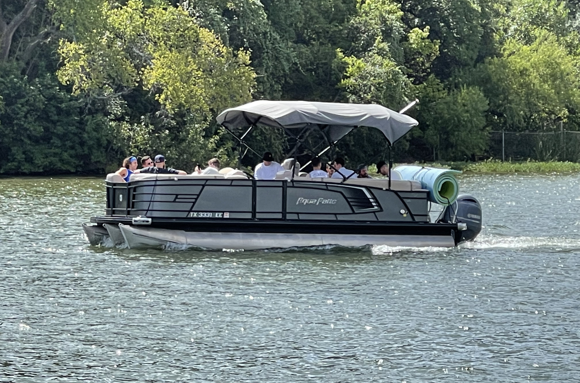 Pontoon boat on water with people; canopy; trees in background.