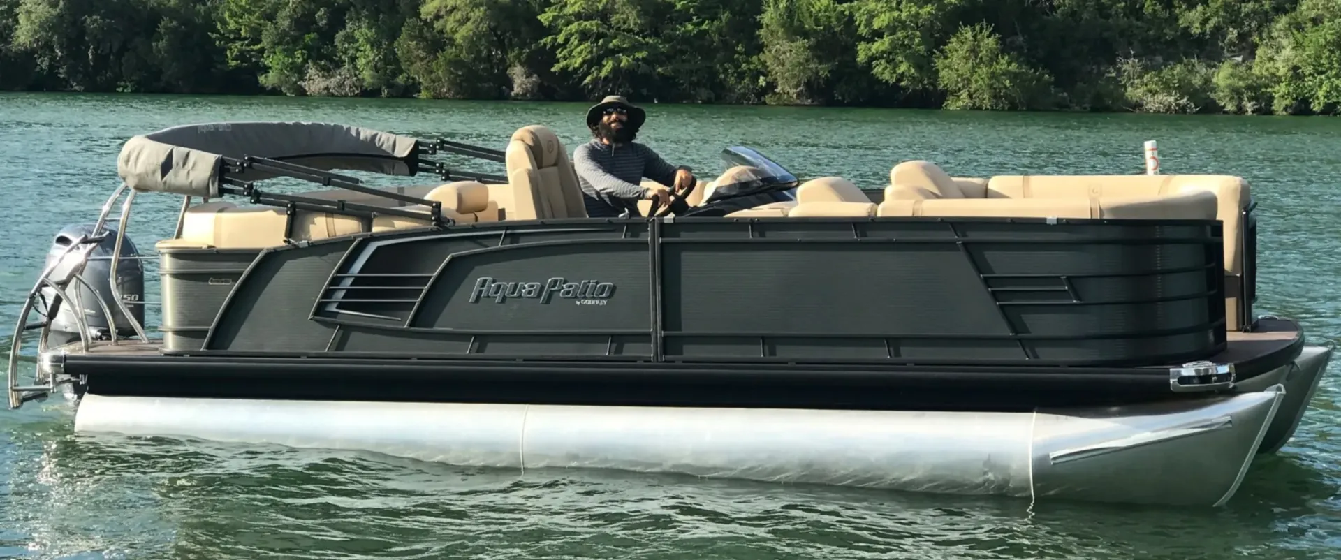 Man piloting a dark green pontoon boat on water, with forested background.