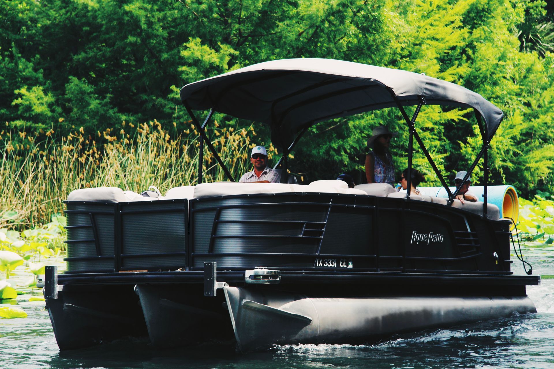 Black pontoon boat with people on it, cruising on a body of water, canopy overhead.