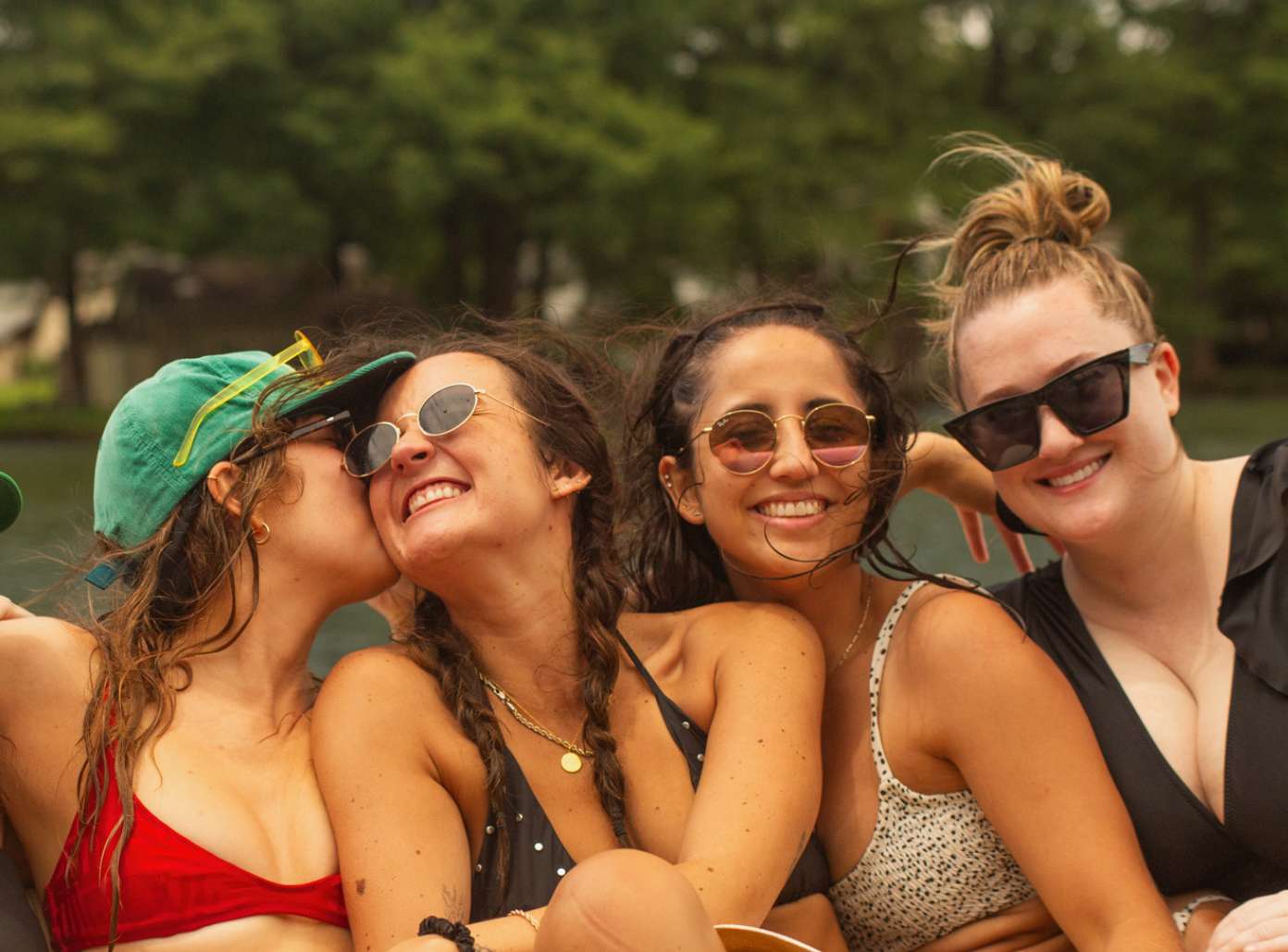 Four smiling friends in swimwear on a boat. One kisses another's cheek. Green trees in the background.