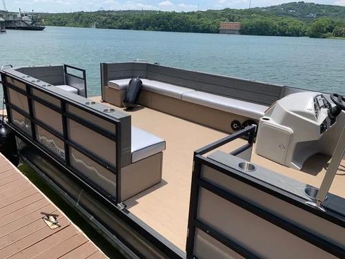 A pontoon boat docked on a lake, featuring gray and tan accents, cushioned seating, and a helm.