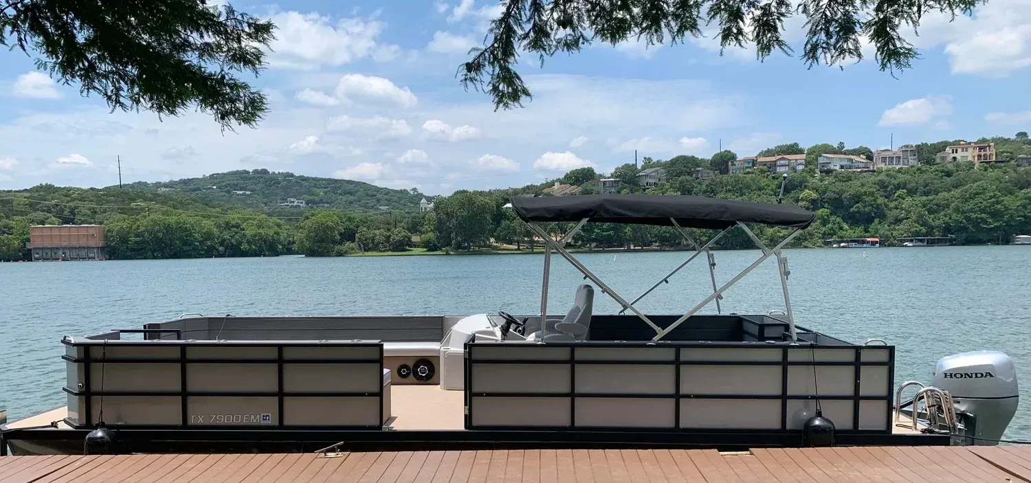 Pontoon boat docked on a wooden pier with a lake and lush green hills in the background on a sunny day.