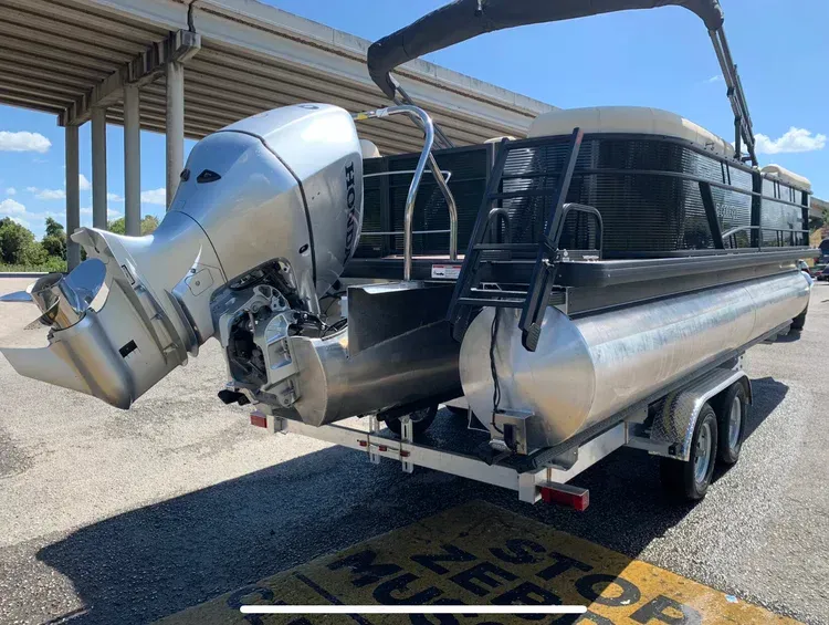 Pontoon boat on trailer, silver motor, parked near a bridge, sunny day.