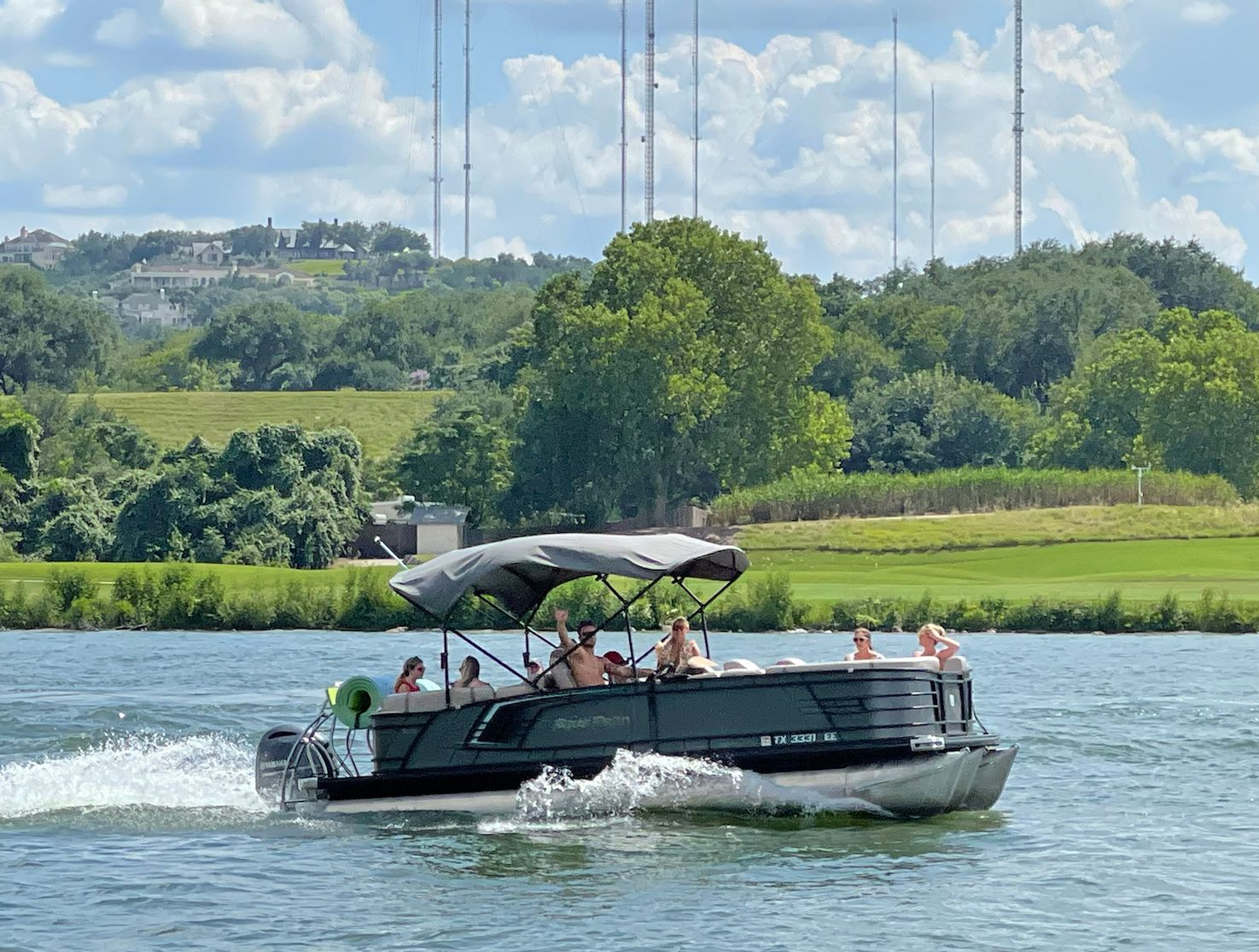 A pontoon boat on a lake with several people aboard, waves, and a green shoreline under a cloudy sky.