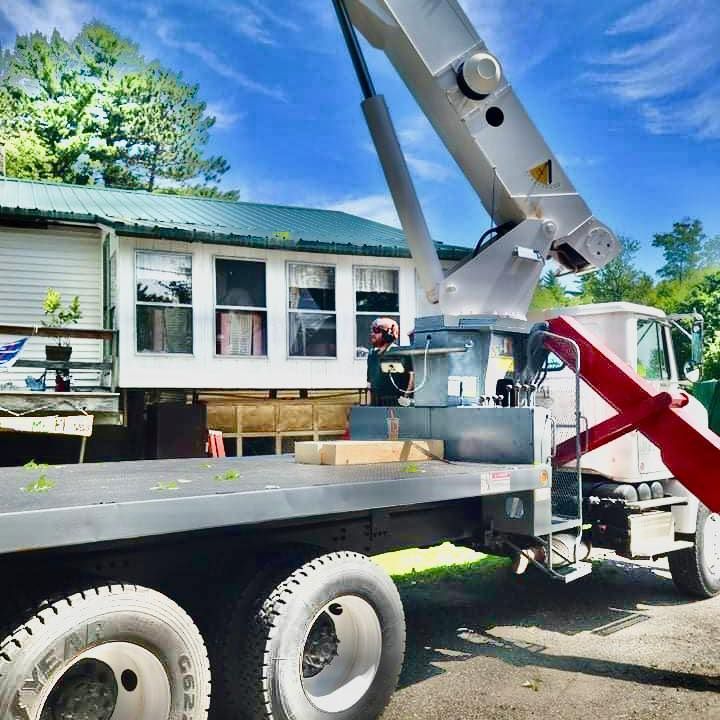 Crane truck lifting a wooden beam near a white house with a green roof on a sunny day.