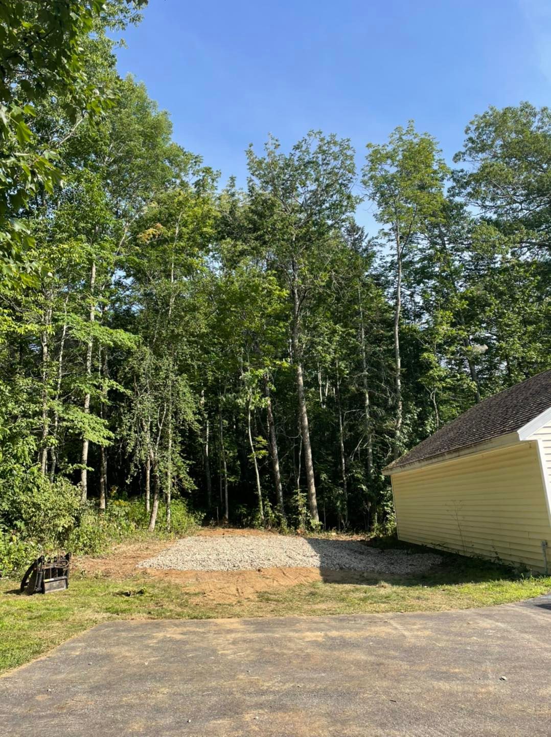 Gravel parking area next to a yellow building and forest under a blue sky.