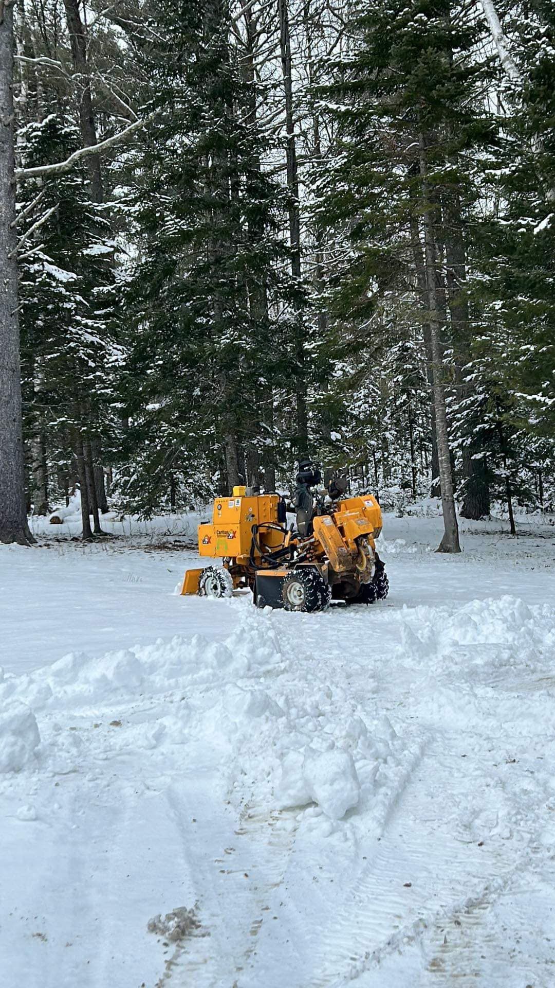 Yellow stump grinder in snow-covered clearing, surrounded by trees.