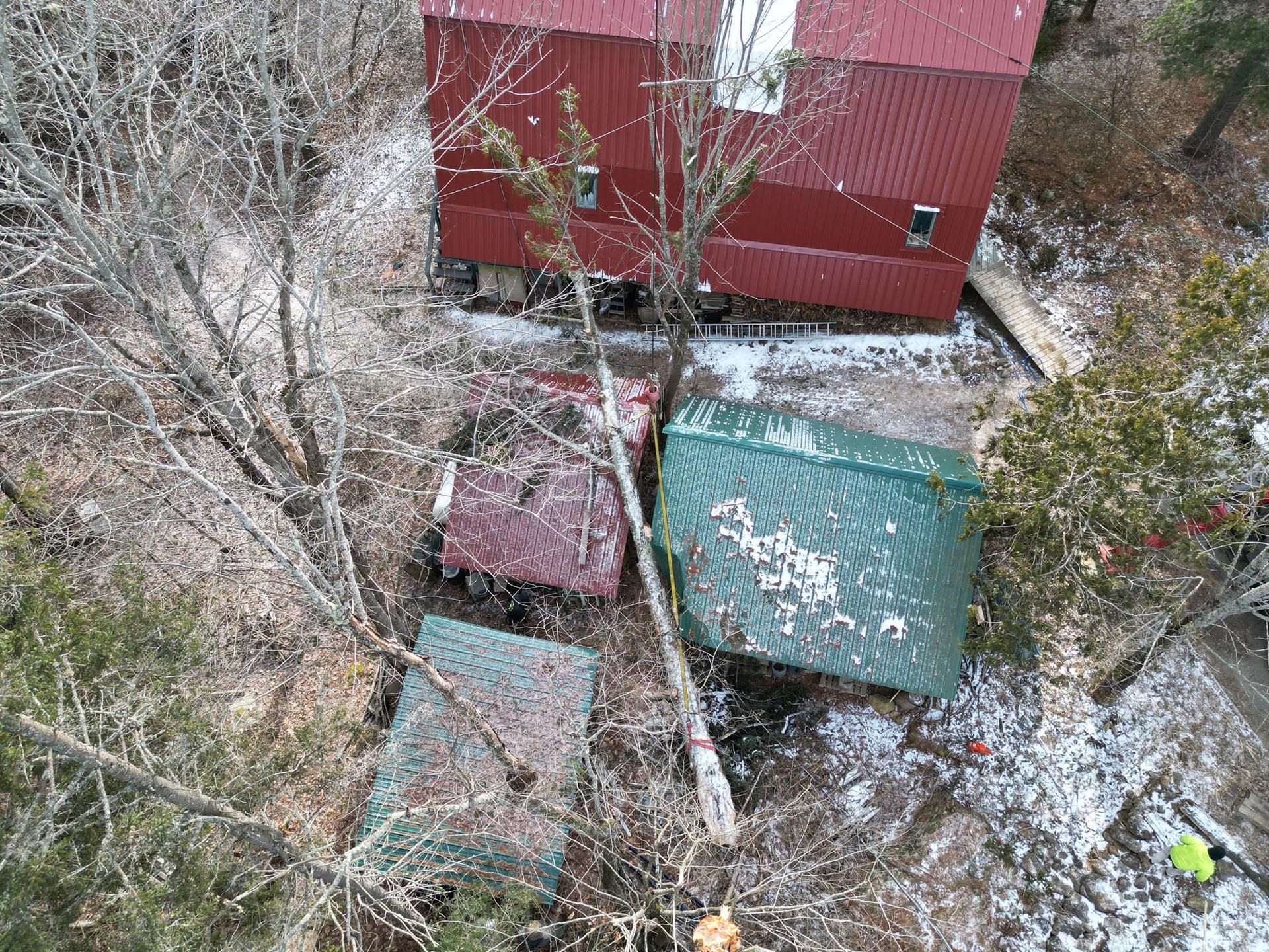 Overhead view of red barn with a tree fallen on two green and red sheds in a snowy woodland.