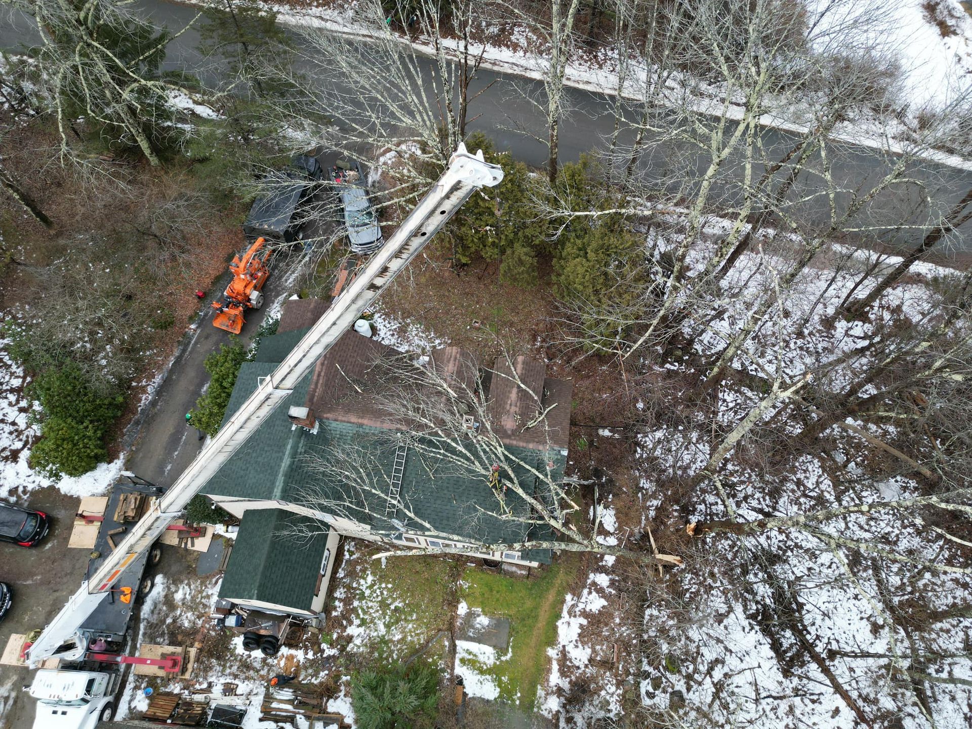 Overhead view of a house with a long crane cutting a tree in a snowy yard, next to a road.