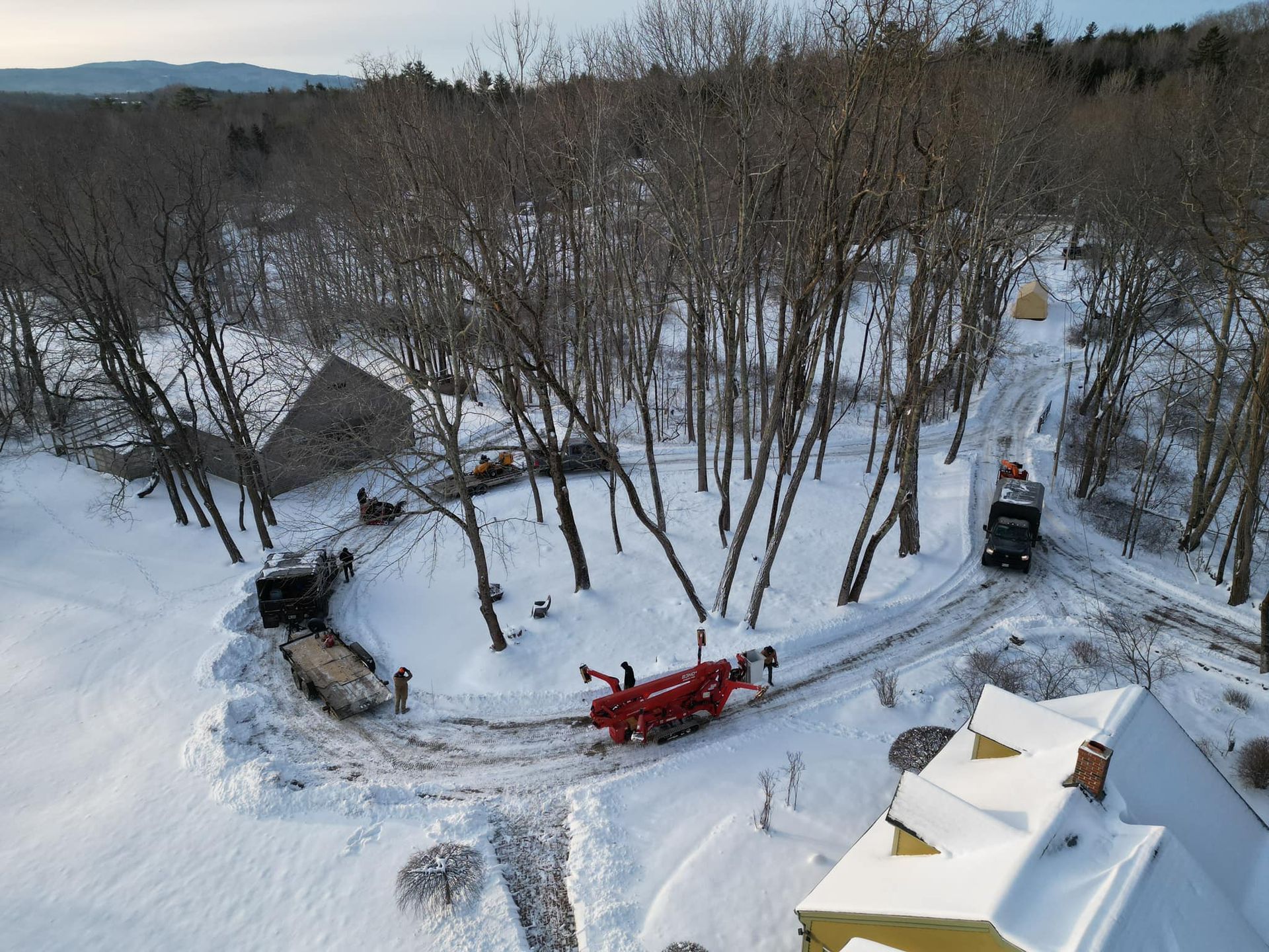 Snow-covered driveway with a snowplow clearing a path. Houses and trees surround the area; winter scene.