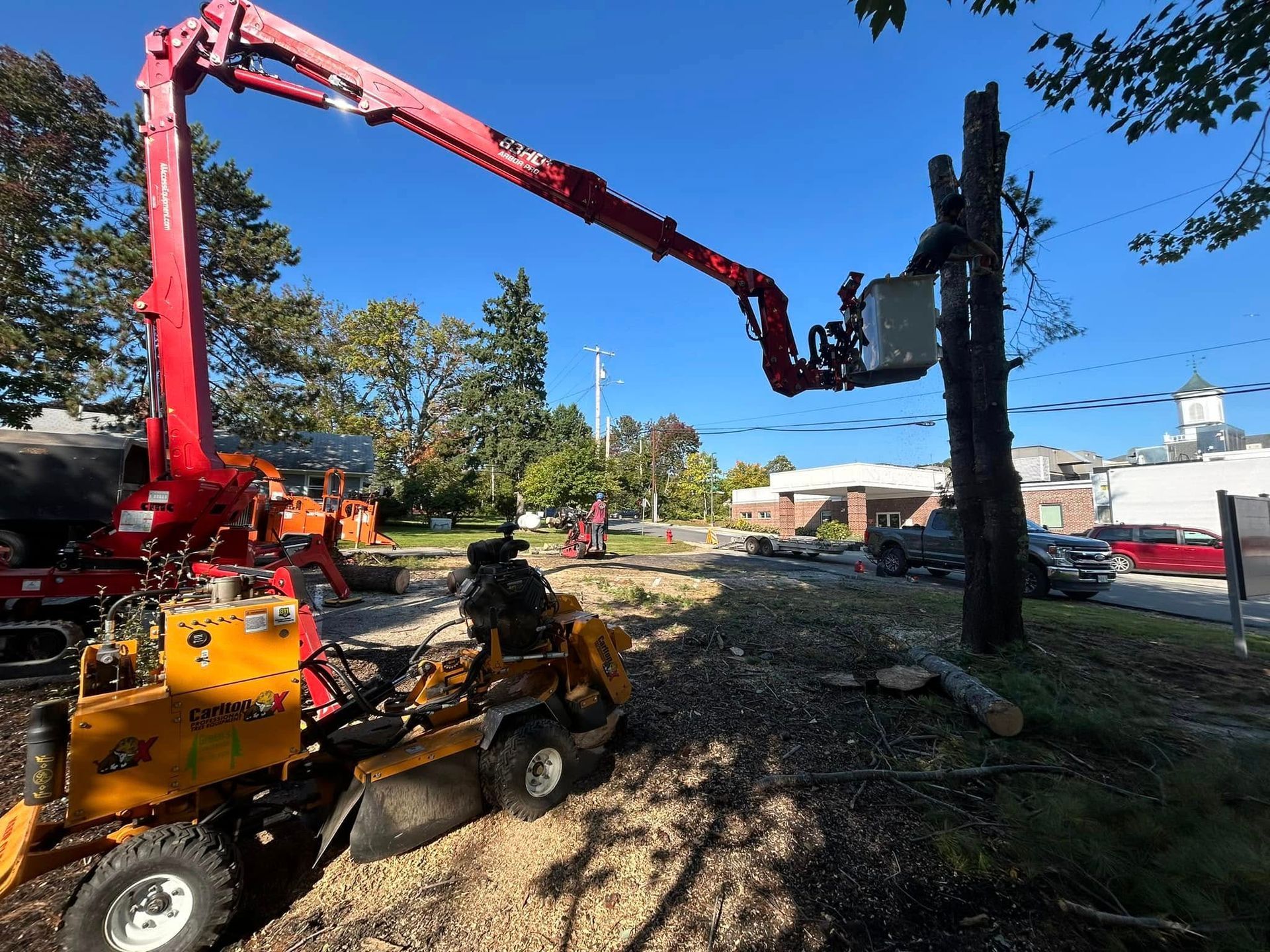 A tree service cutting a tree with a cherry picker and a wood chipper in a grassy area.