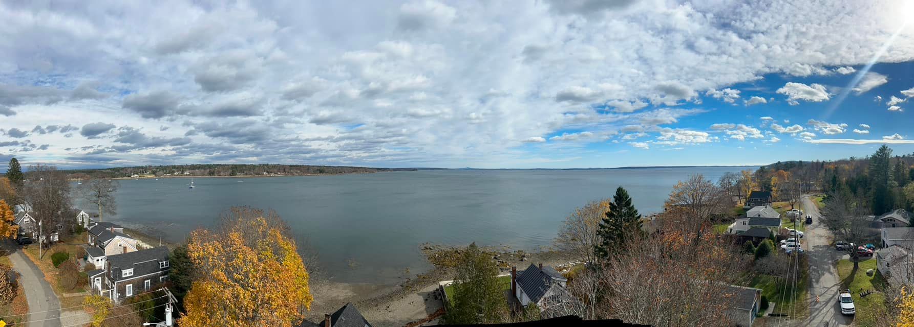 Panoramic view of a body of water with a cloudy sky, houses, and trees in fall colors.