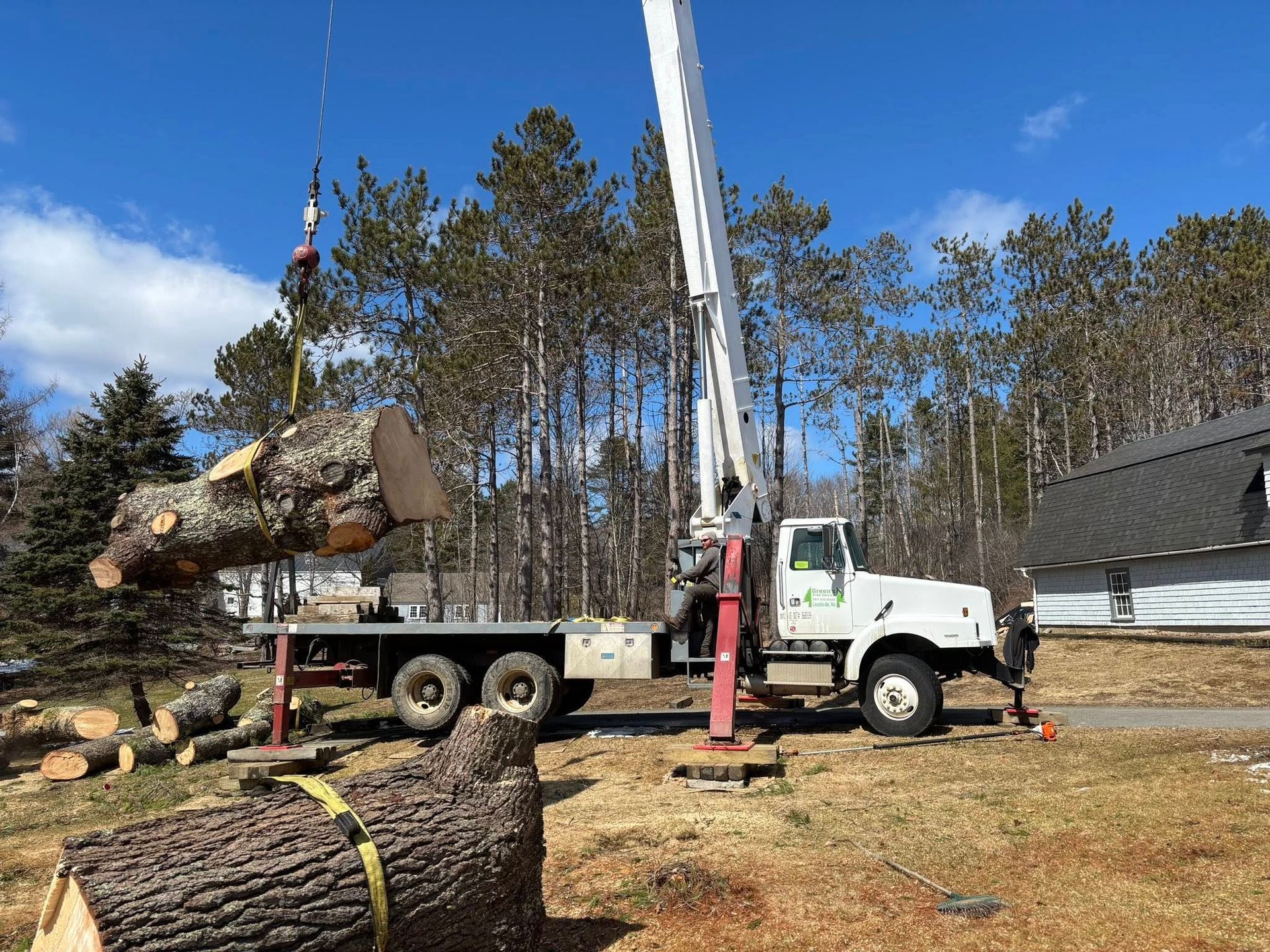 A crane lifts a large tree trunk onto a flatbed truck in a residential yard.