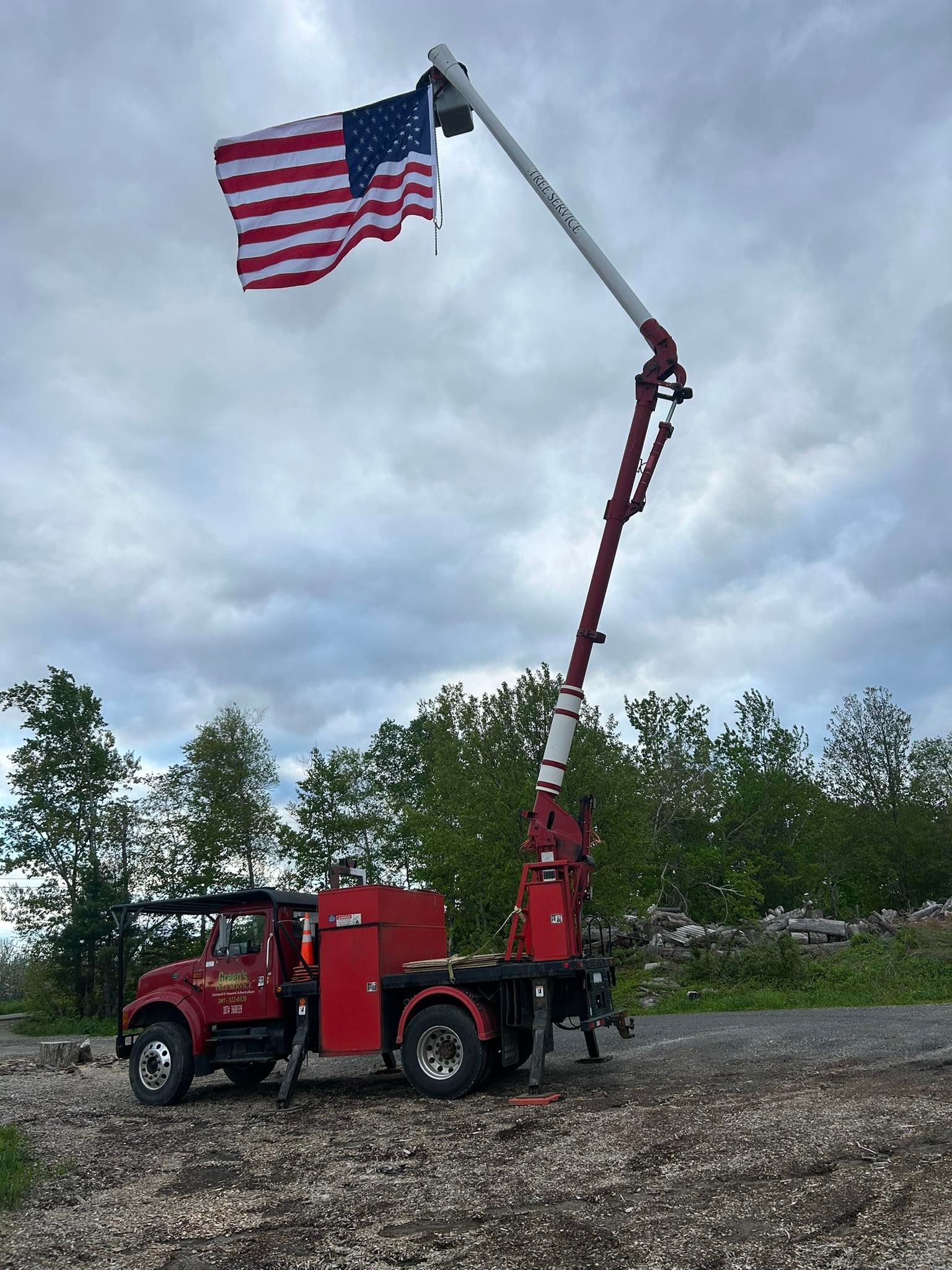 Red truck with extended crane arm, American flag flies at the end. Cloudy sky, trees in background.