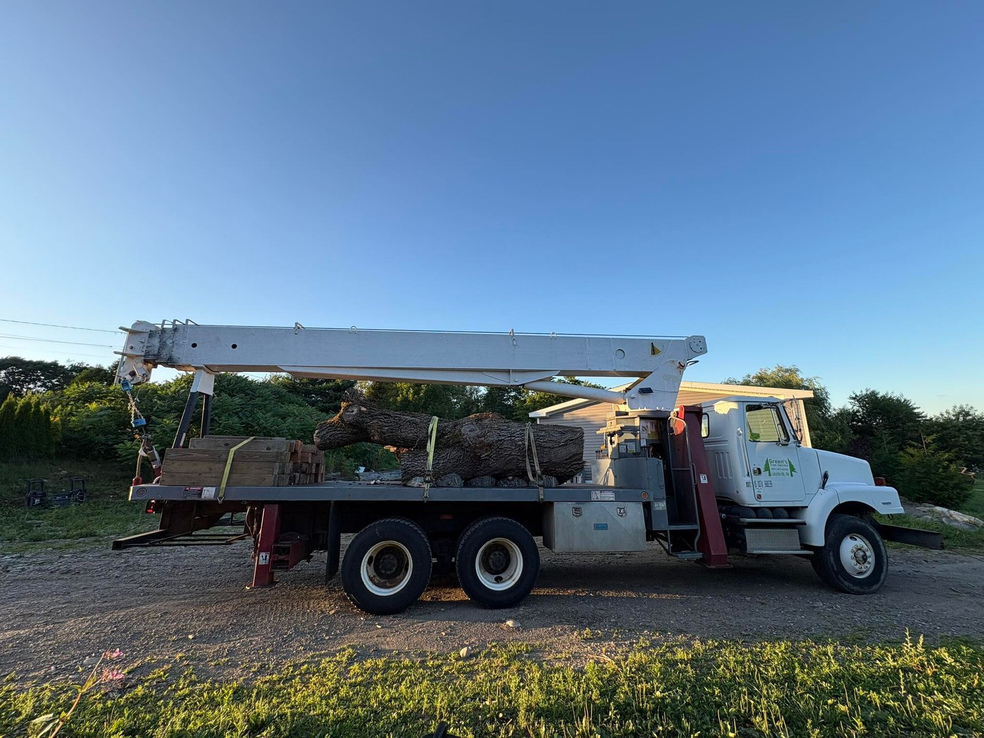 White tree service truck with a raised crane boom, loaded with logs, parked on gravel.