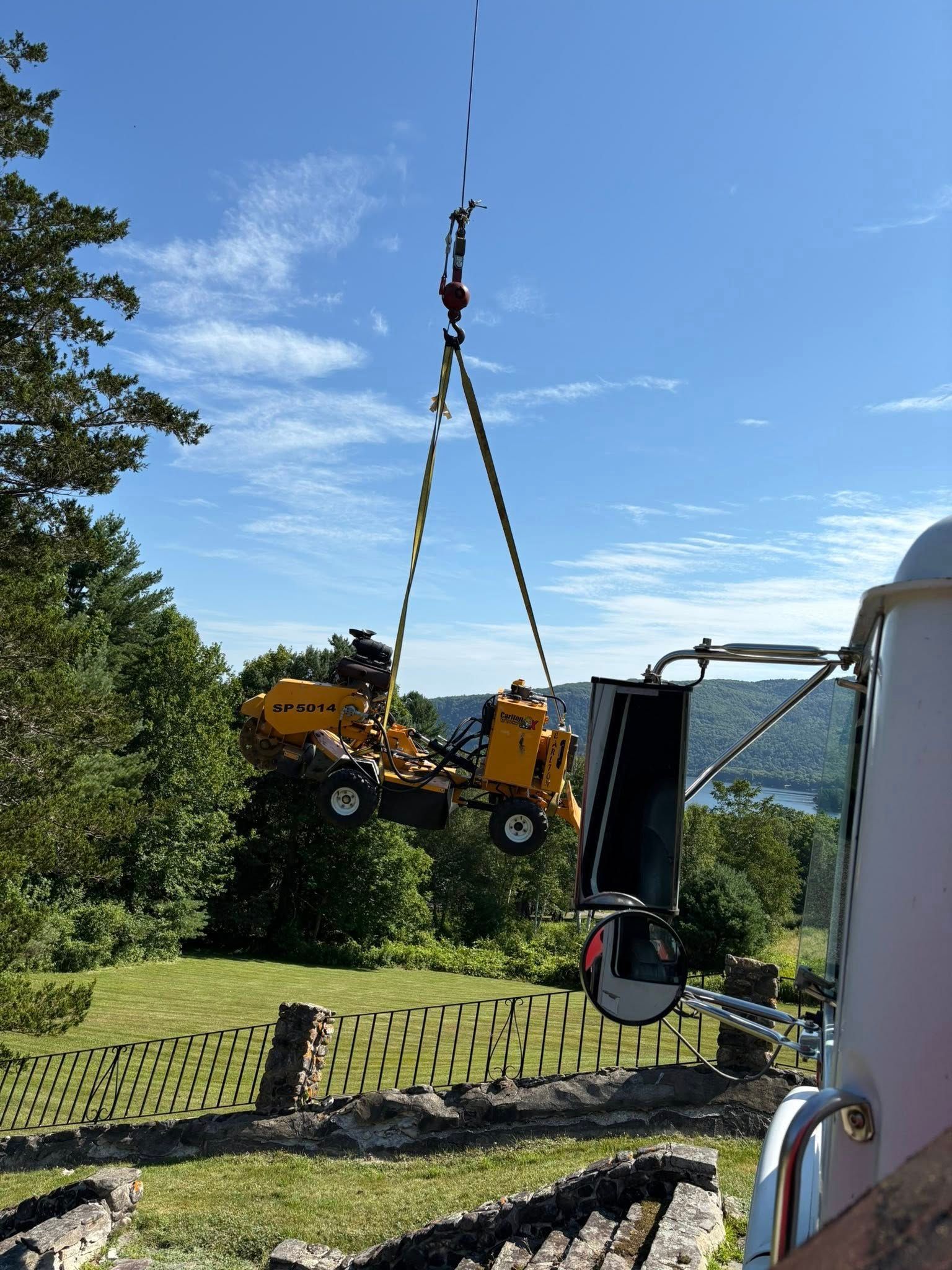 A yellow construction machine is lifted by a crane in a green, outdoor setting.