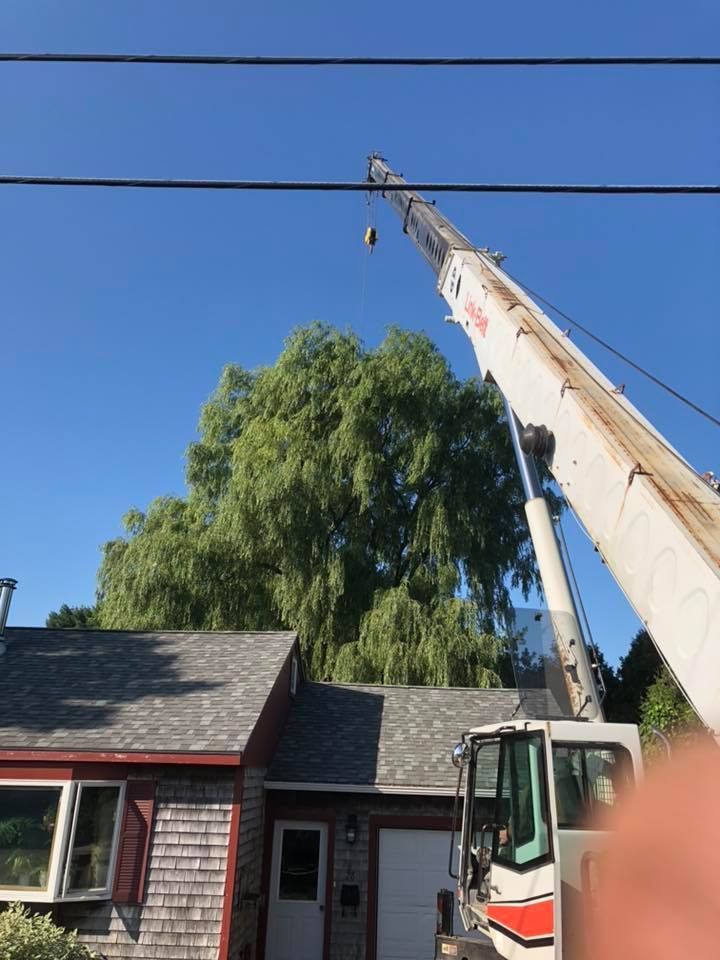 A crane reaching for electrical wires above a house, tree, and blue sky.
