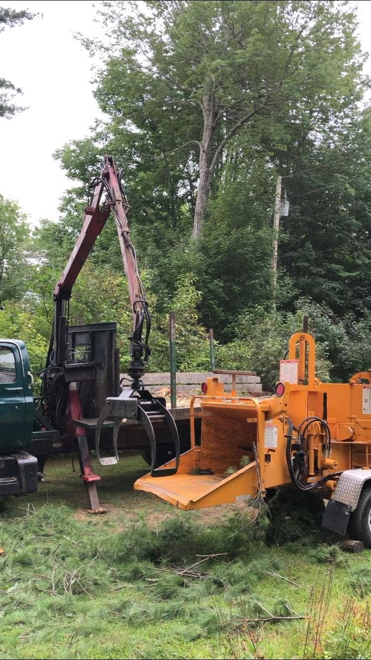 A truck with a grapple arm feeds a wood chipper in a grassy area, trees in the background.