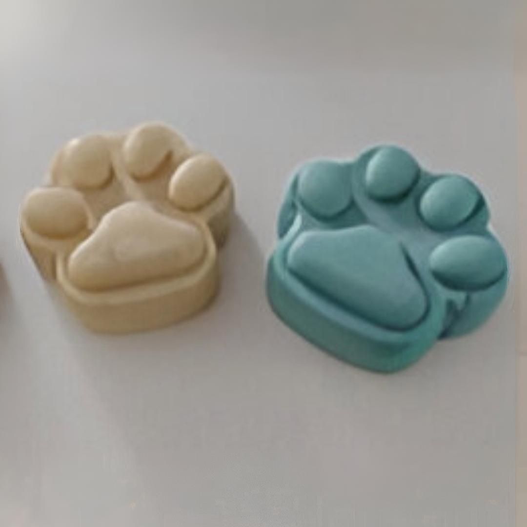 Dos jabones con forma de pata, uno de color crema y otro azul, apoyados sobre una superficie blanca.