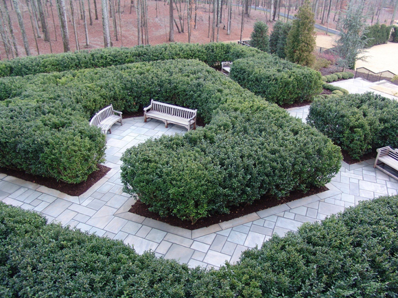 Top-down view of a garden maze with green hedges, stone pathways, and benches.